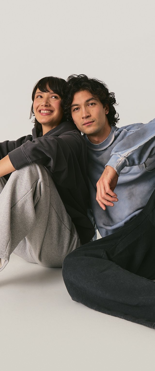 Two individuals sit together, wearing a dark gray oversized hoodie and a light blue sweatshirt with distressed edges, paired with gray and black joggers.