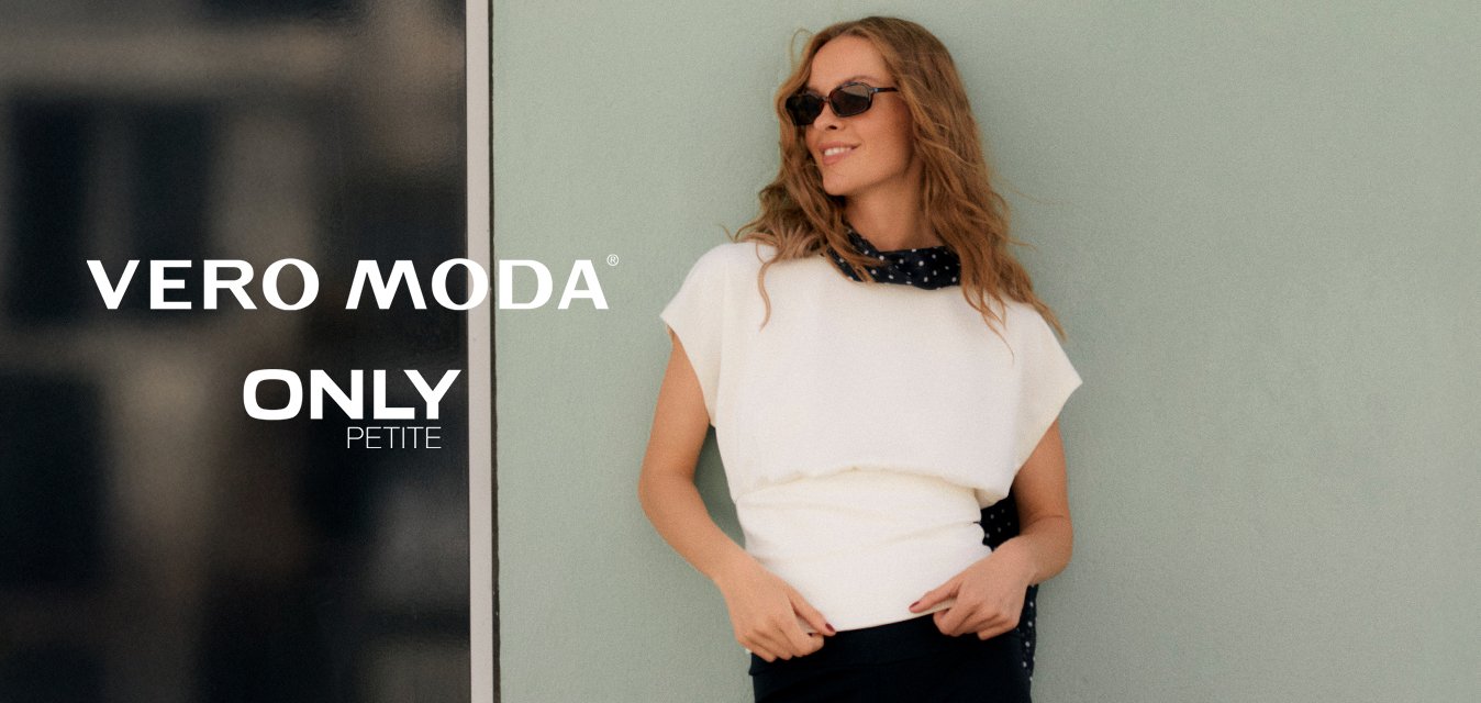 Žena s vlnitými vlasy, která má na sobě sluneční brýle, bílý top, černé tříčtvrteční kalhoty a černý šátek s bílými puntíky, pózuje u světle zelené zdi.