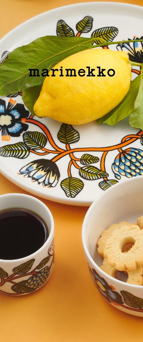 Limón con hojas verdes en un plato con estampado floral, taza de café negro y bol con galletas redondas de mantequilla sobre superficie naranja.