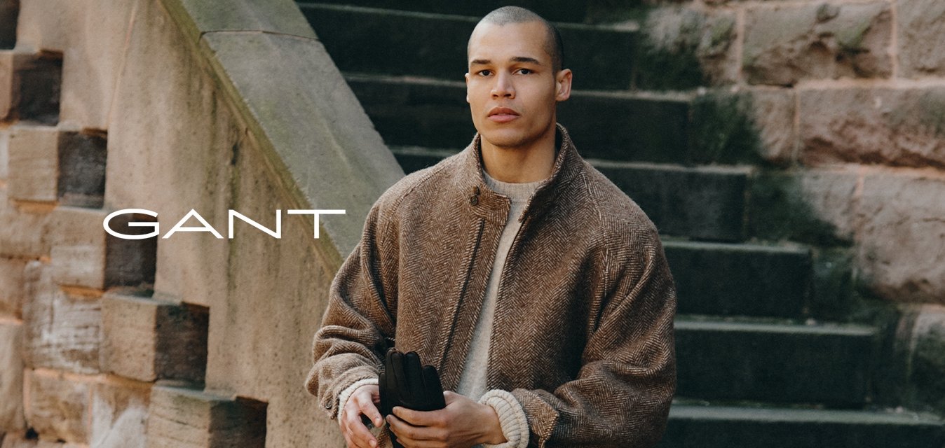 Hnědá texturovaná bomber bunda přes krémový pletený svetr, tmavé džíny a černé boty. Stojí na kamenitých schodech, přičemž je vidět cestu.