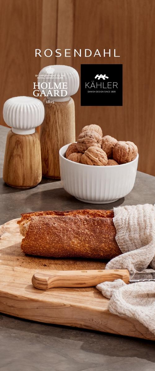 Trækværne til salt og peber, hvid skål med valnødder, delvist skåret baguette med serrateret kniv på træbræt med klud.