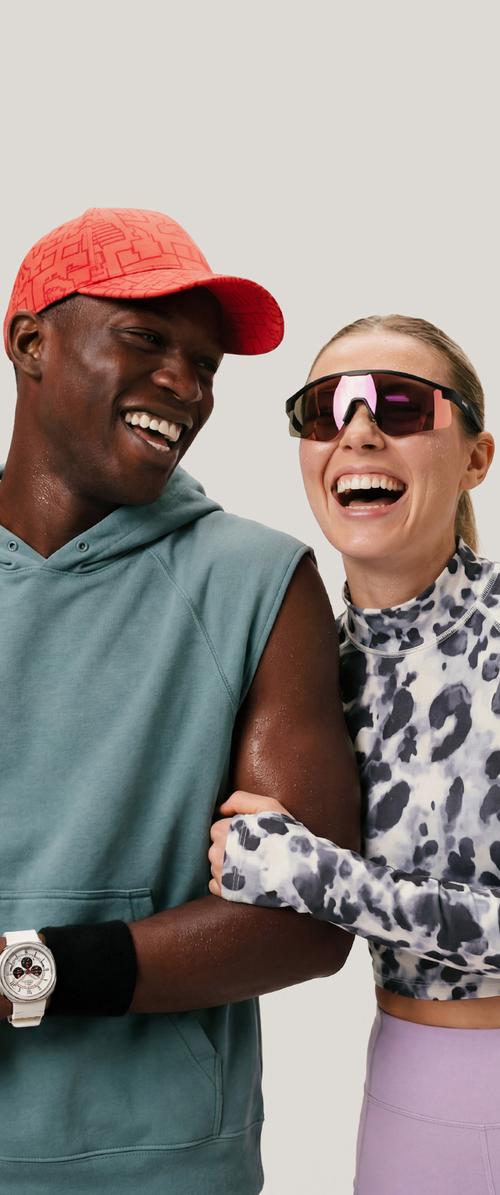 Zwei Personen in Sportbekleidung lächeln und lachen, eine trägt eine rote Kappe und ein ärmelloses Hoodie, die andere große Sportsonnenbrillen.