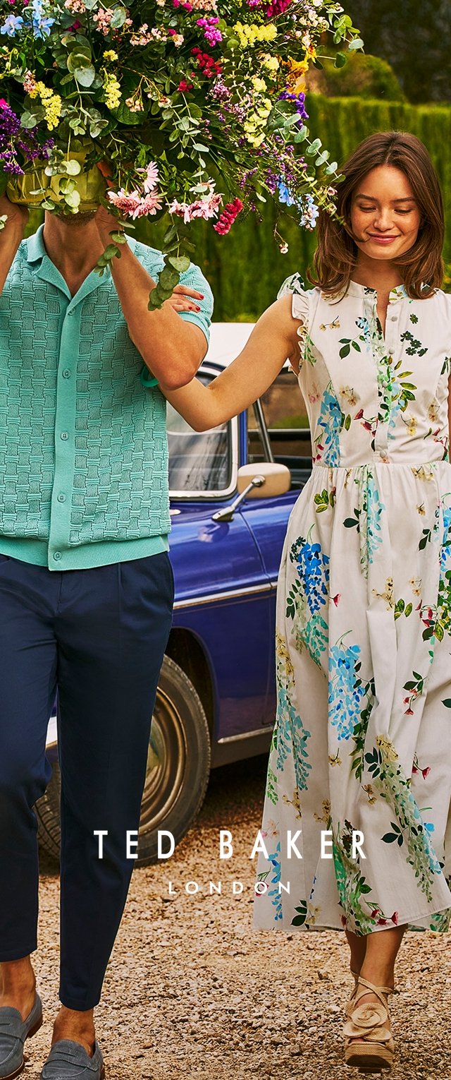 Turquoise gebreide cardigan over een witte jurk met bloemenmotieven in blauw en groen; beide dragen marineblauwe broeken en de vrouw heeft beige sandalen aan.