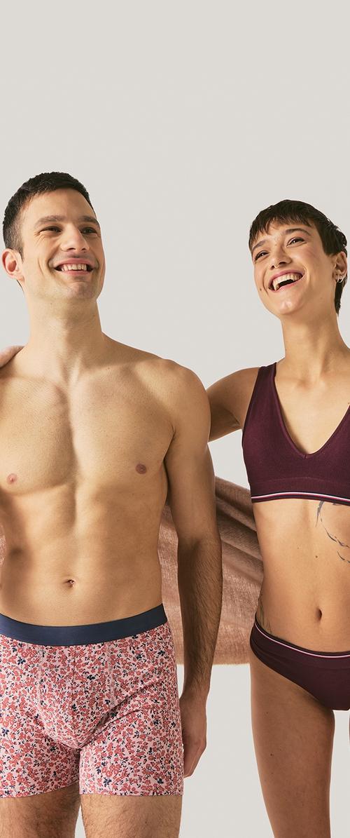 Glimlachende man en vrouw in ondergoed staan naast elkaar; de man draagt boxershorts met bloemmotief, de vrouw een bijpassende bordeauxrode beha en slip.