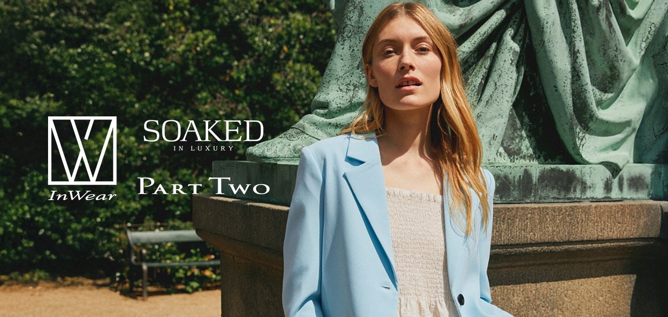 Žena s dlouhými blonďatými vlasy, oblečená v světle modrém saku a bílých šatech, stojí blízko velkého zeleného soklu sochy venku.