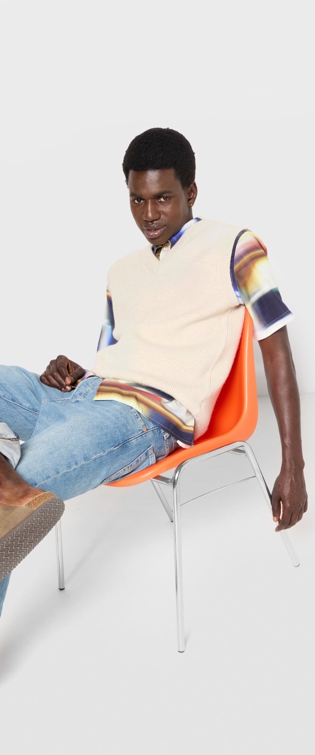Chaleco de punto crema sobre una camiseta de manga corta multicolor, combinado con pantalones vaqueros de mezclilla azul claro. Sentado en una silla naranja con patas de cromo.