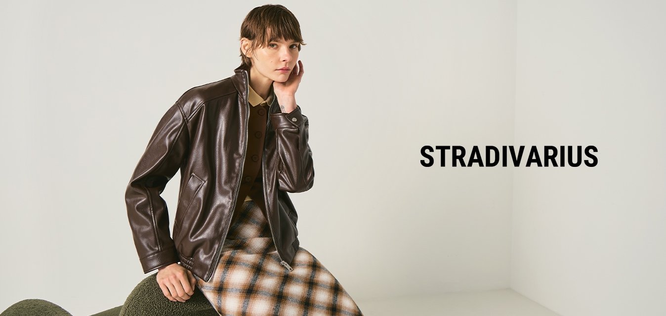 Osoba v tmavě hnědém koženém kabátu a kostkované sukni, sedící na zelené texturované židli na jednobarevném světlém pozadí.