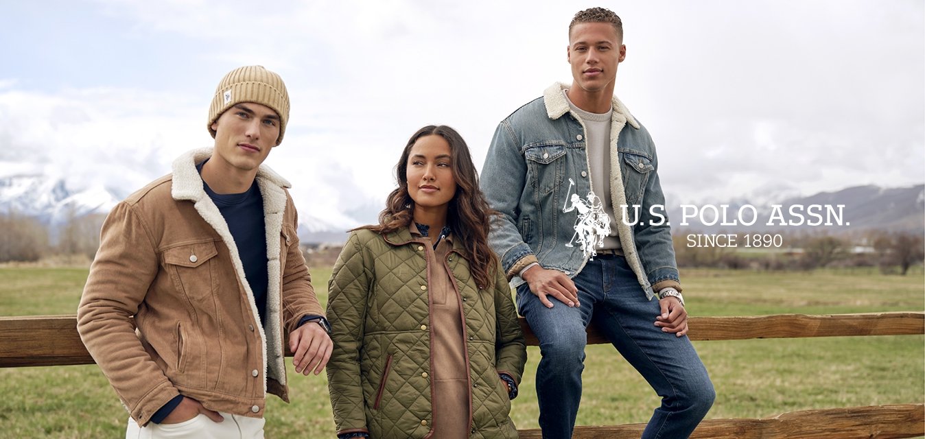 Tři mladí dospělí v zimních bundách a botách pózují u dřevěného plotu na travnatém poli s zasněženými horami v pozadí.