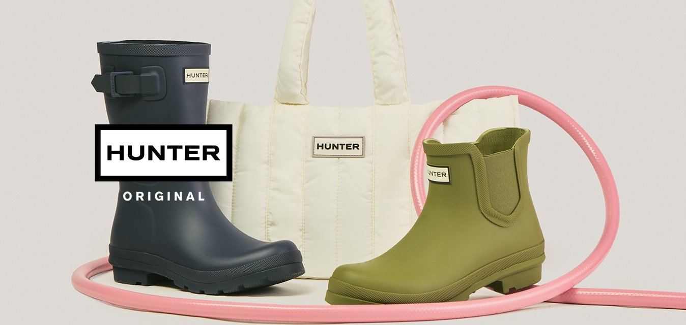 Vysoká námořnicky modrá dešťová bota, krátká kotníková bota olivově zelené barvy, bílá prošívaná taška přes rameno, všechny s logem Hunter, uspořádané vedle stočené růžové hadice.