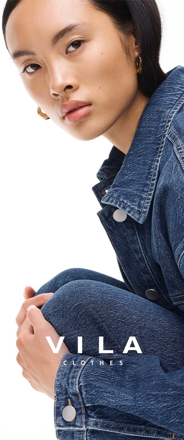 Giovane donna con capelli corti e scuri, che indossa orecchini a cerchio dorati e una giacca di jeans blu, seduta con le mani intrecciate attorno al ginocchio.