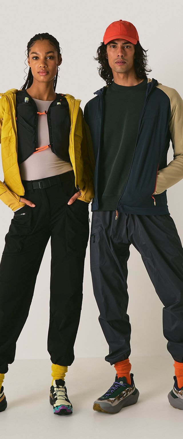 Zwei Models tragen Sportbekleidung: eine gelbe Daunenjacke, schwarze Hosen, ein dunkelgrünes Shirt und bunte Sneaker mit sichtbaren Texturen und Akzenten.