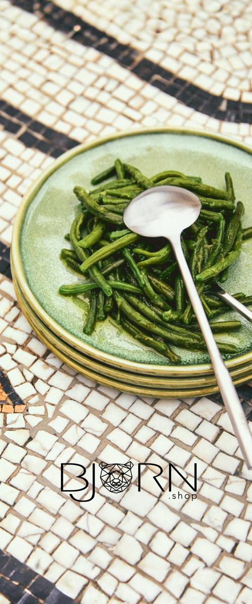 Des haricots verts sont disposés sur une assiette verte texturée, accompagnés d'une cuillère en argent brillante. L'arrière-plan présente un motif en mosaïque.