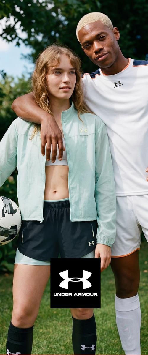 Deux joueurs de football en tenue de sport Under Armour se tiennent ensemble à l’extérieur ; l’un tient un ballon de football, tous deux semblent détendus et confiants.