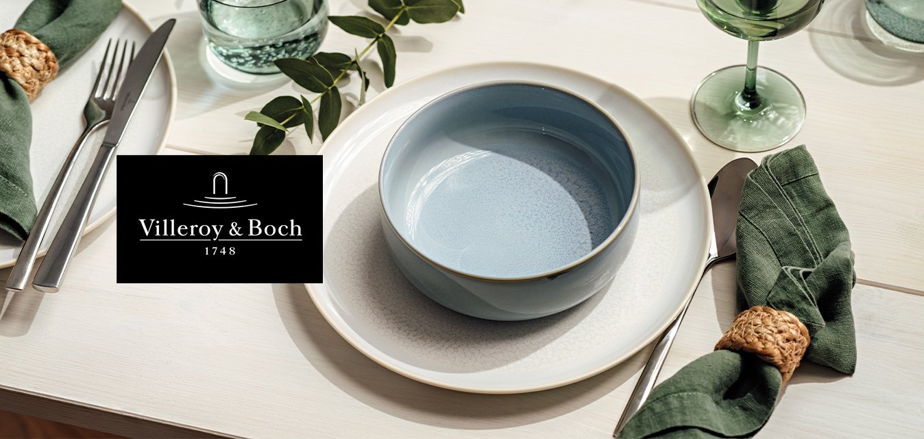 Bílé jídelní talíře s modrou miskou, zelené ubrousky v tkaných kroužcích, příbory, čiré a zelené sklenice a dekorace se zelenými listy na stole.