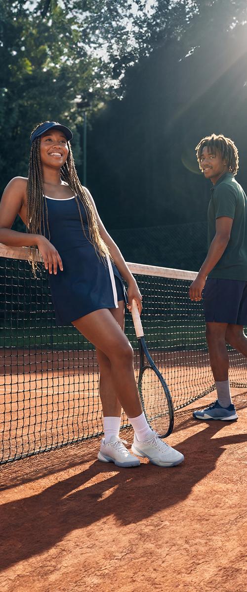 Zwei junge Erwachsene in Tenniskleidung stehen an einem Netz auf einem Außen-Tonplatz mit Bäumen im hellen Sonnenlicht.