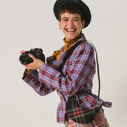 Mladá žena v kostkovaném oblečení a černých botách drží fotoaparát a hravě pózuje s jednou nohou zvednutou na jednobarevném pozadí.
