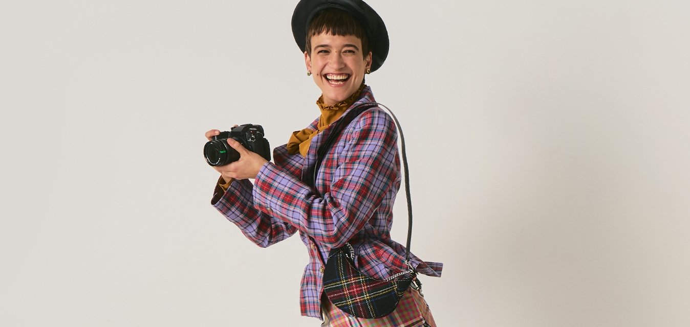 Mladá žena v kostkovaném oblečení a černých botách drží fotoaparát a hravě pózuje s jednou nohou zvednutou na jednobarevném pozadí.