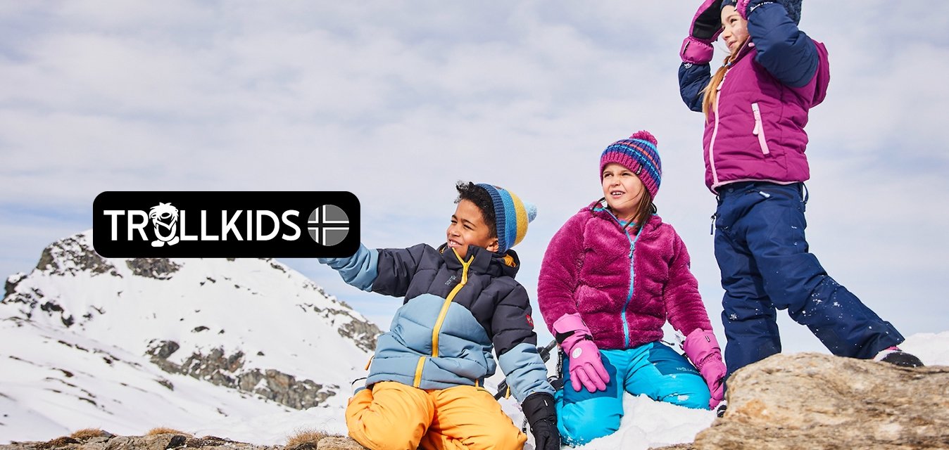 Tři děti v barevných zimních bundách: modré a šedé, fialové a námořnické. Sedí na kameni v zasněžené krajině s horami v pozadí.