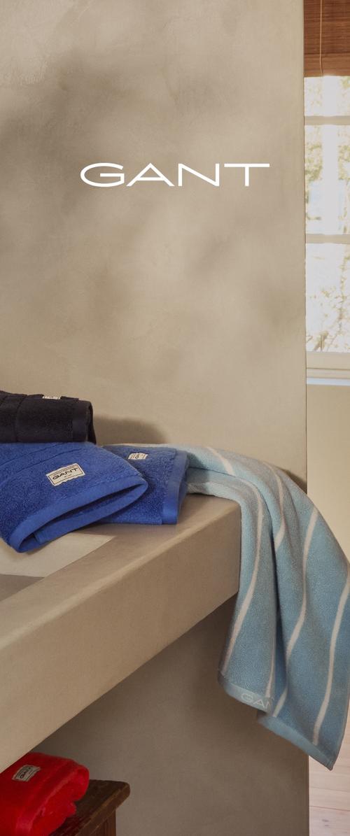 Serviettes bleues et bleu marine avec une texture douce et des étiquettes GANT, drapées sur une étagère beige. Une serviette présente un motif de rayures bleu clair.
