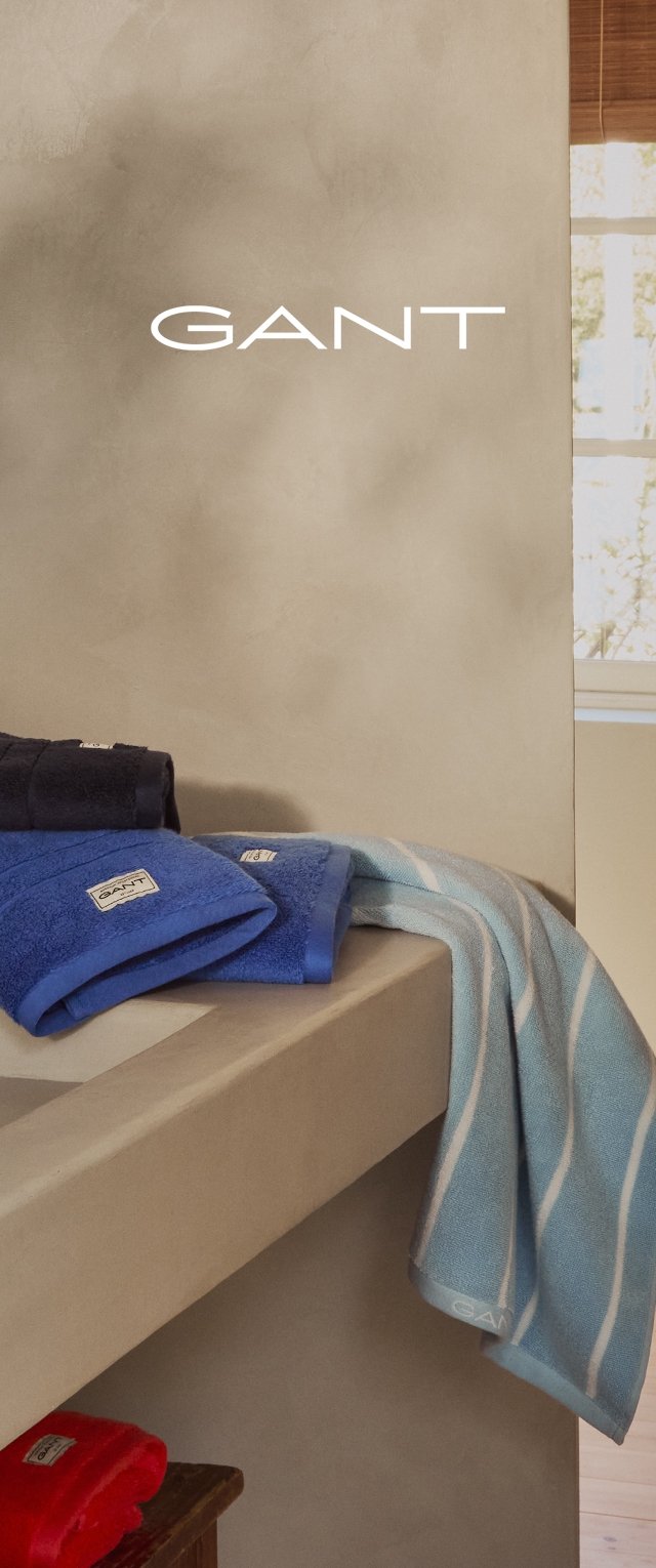Toallas en tonos azul y marino con una textura suave y etiquetas de GANT, cayendo sobre una estantería beige. Una de las toallas presenta un patrón de rayas en azul claro.