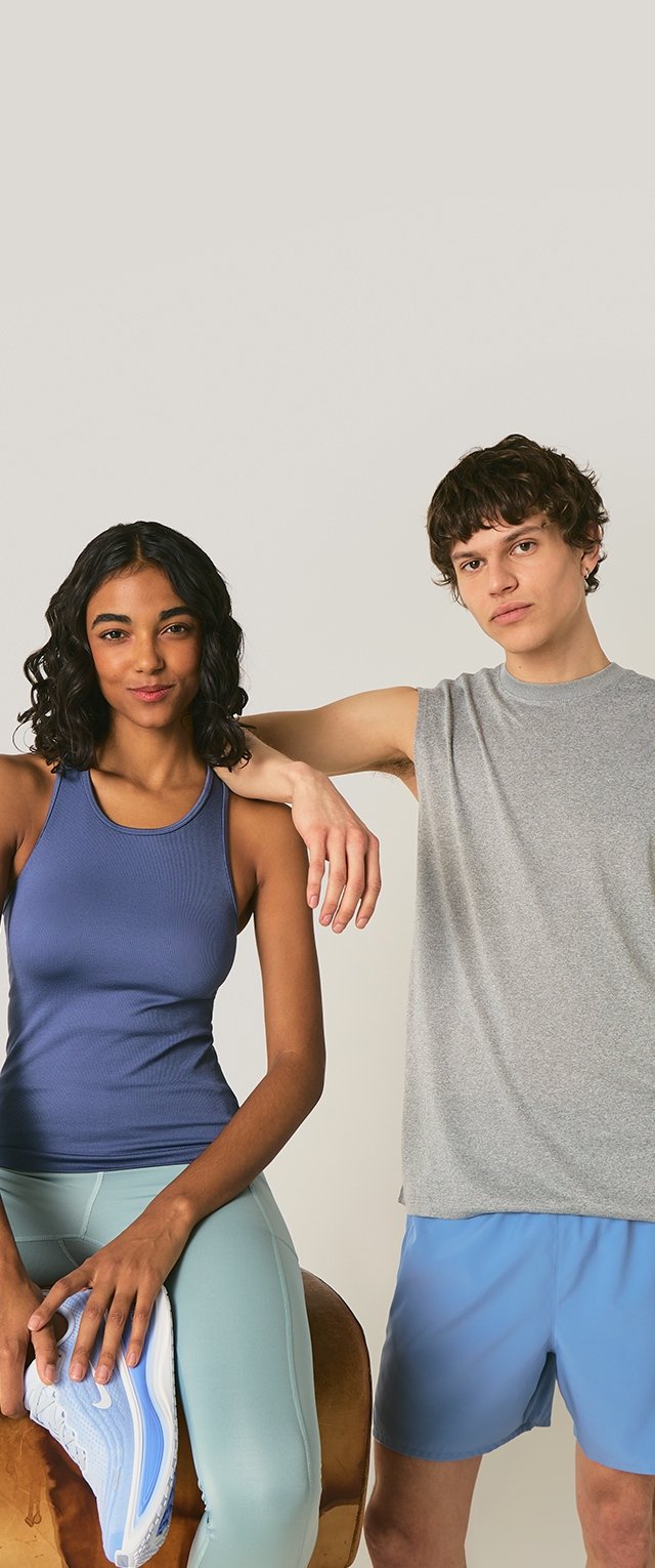 Hombre con camiseta sin mangas gris y pantalones cortos azules, de pie con el brazo sobre el hombro de una mujer; mujer con camiseta sin mangas morada y mallas azul claro sostiene una zapatilla.