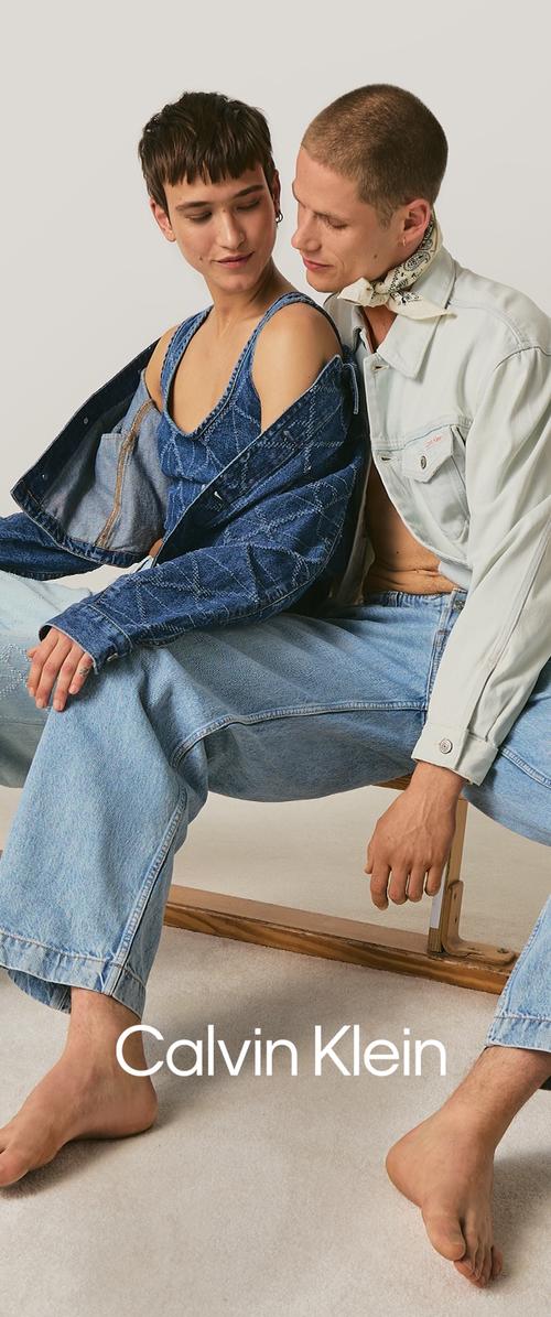 Dos personas sentadas cerca, con chaquetas ligeras y prendas de mezclilla, posando descalzas y relajadas frente a un fondo liso.