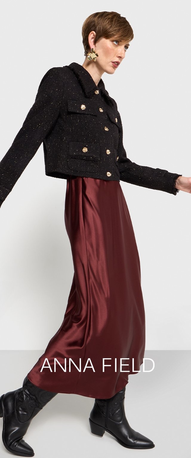Čierna textúrovaná skrátená bunda so zlatými gombíkmi, nosená cez burgundskú saténovú maxi šaty, doplnená s vysokými čiernymi kovbojskými čižmami.
