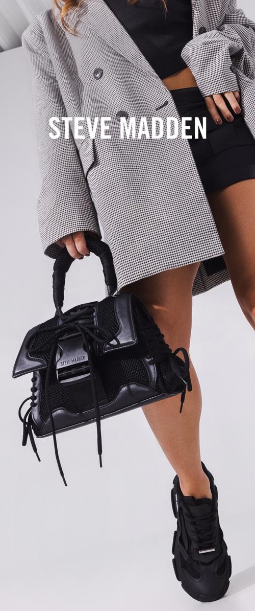 Kvinna som bär svarta shorts, hundtandsmönstrad blazer och svarta sneakers, håller en svart Steve Madden-handväska i handtagen.