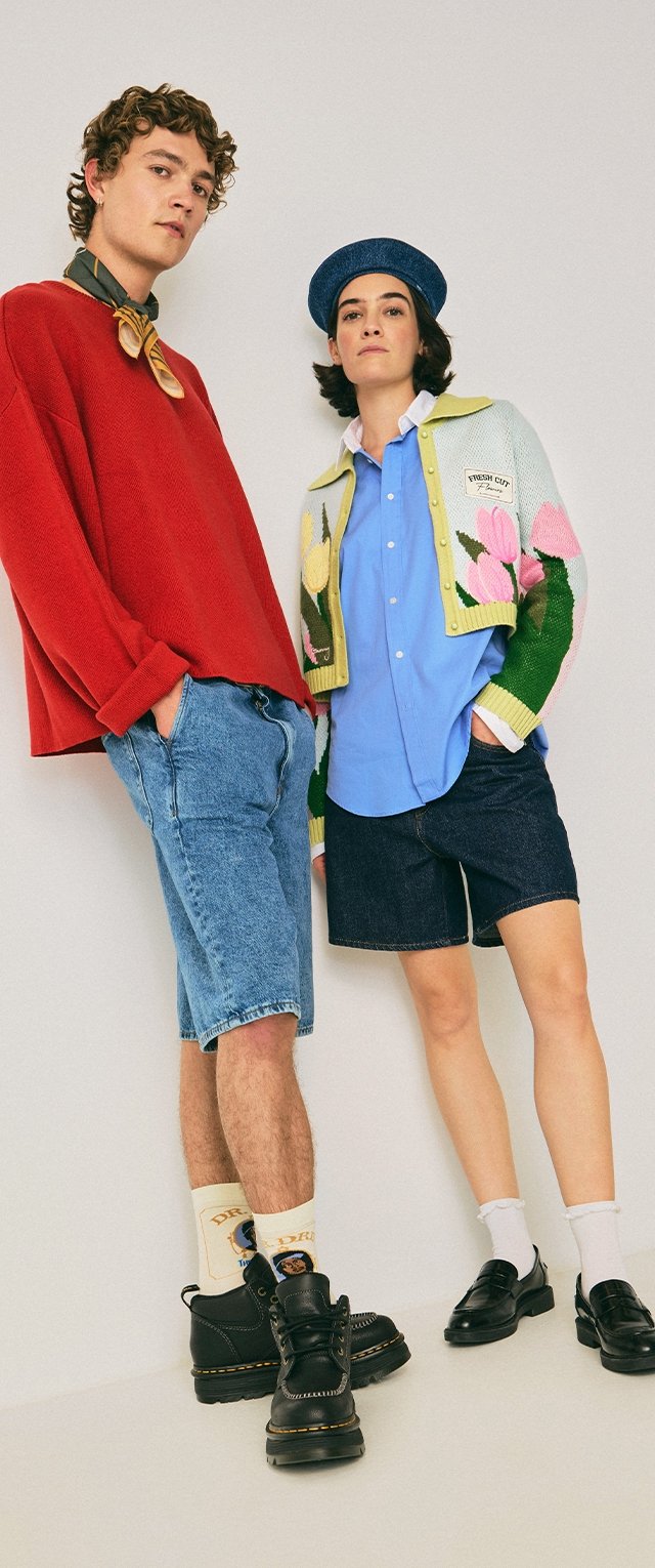 Dos personas de pie, una con un suéter rojo, pantalones cortos de mezclilla y botas negras, y la otra con una camisa azul, un cárdigan floral, boina y mocasines negros.