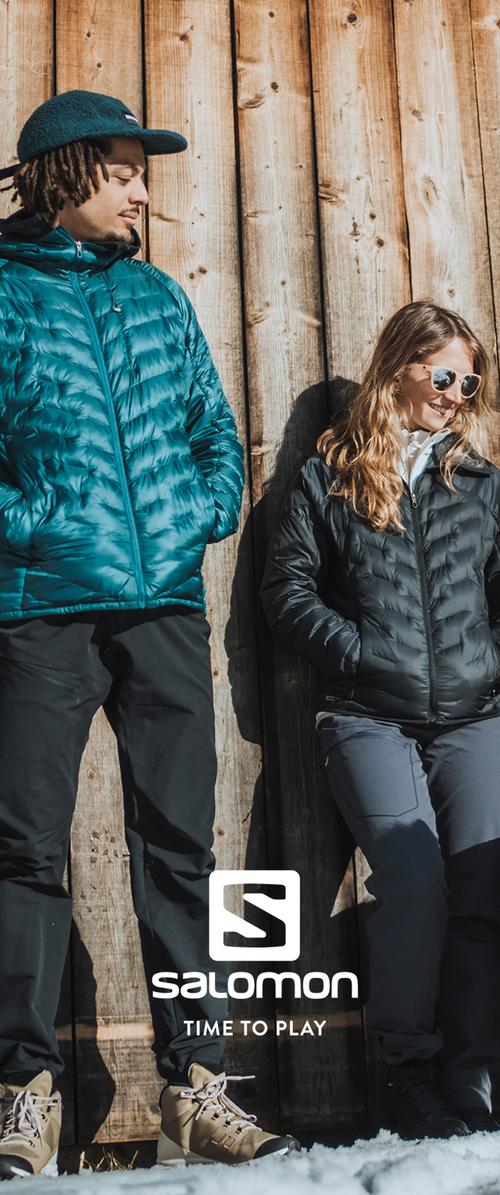 Dos personas están de pie contra una pared de madera. Una lleva una chaqueta acolchada azul verdoso, pantalones negros y una gorra. La otra lleva una chaqueta acolchada negra y pantalones grises.