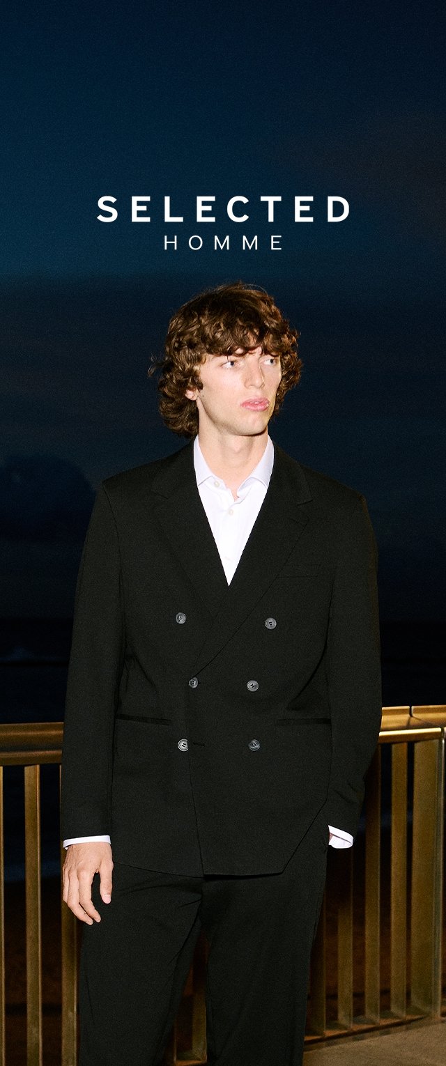 Jonge man met krullend haar in een zwart dubbelgeknopt pak en een wit overhemd, staande bij een reling 's nachts onder de tekst "SELECTED HOMME".