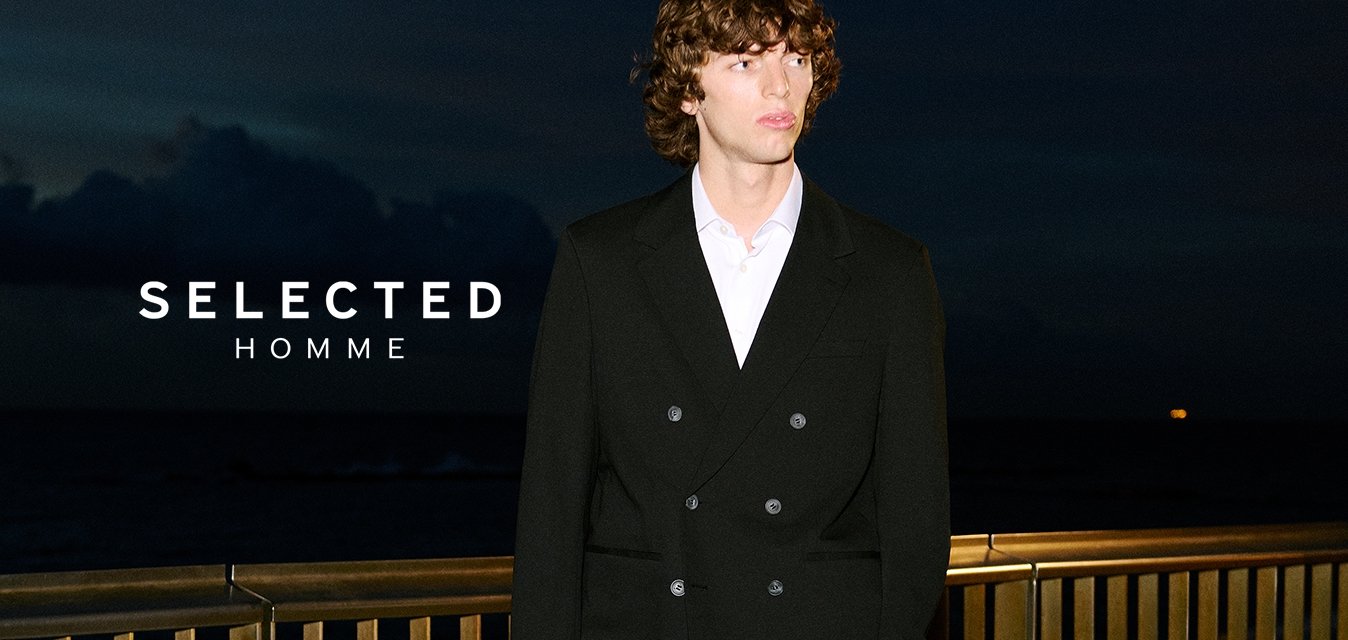 Mladý muž v černém dvouřadém obleku a bílé košili stojící na kamenné terase u kovového zábradlí v noci.