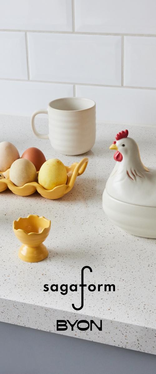 Vitt bänkskiva med en vit keramikkopp, ett gult äggställ med blandade ägg, en behållare formad som en höna och en liten gul äggkopp.