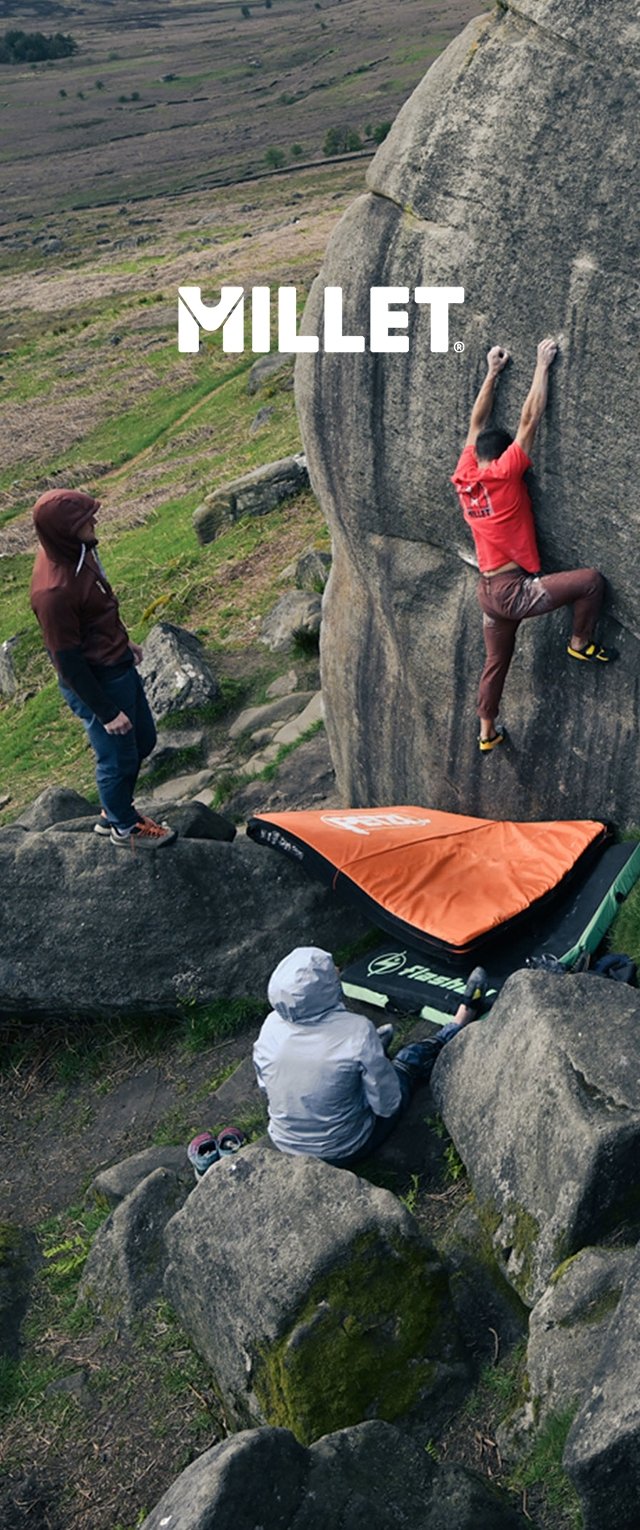 Escena de escalada en roca con un boulder gris. Un escalador vestido de rojo se estira hacia arriba, mientras que dos espectadores con chaquetas están cerca. Una colchoneta de impacto de color naranja brillante está situada abajo.