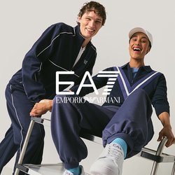 Dva mladí muži v tmavě modrých teplákových soupravách se usmívají, jeden tlačí druhého, který sedí na kovovém vozíku, na jednobarevném pozadí.