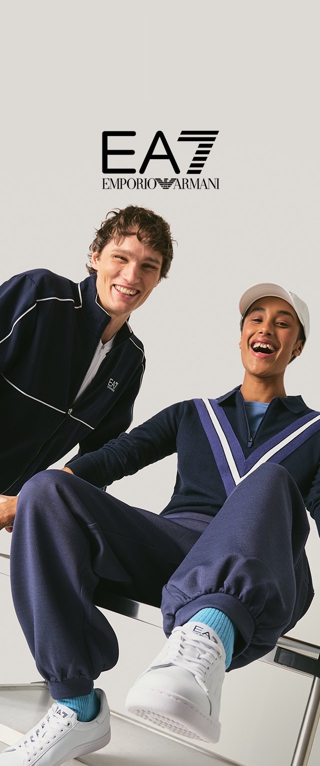 Dos modelos sonrientes con trajes deportivos azul marino y zapatillas blancas posan bajo el logo de EA7 Emporio Armani sobre un fondo claro.