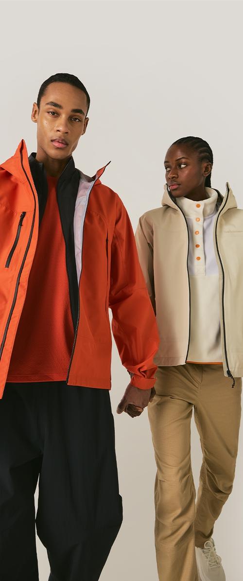Mies oranssissa takissa ja mustissa housuissa pitää kädestä naista, jolla on beige takki, valkoinen paita ja khakinväriset housut yksiväristä taustaa vasten.