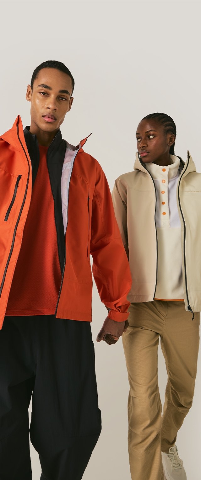 Hombre con chaqueta naranja y pantalones negros tomados de la mano con una mujer con chaqueta beige, camiseta blanca y pantalones caqui sobre un fondo liso.