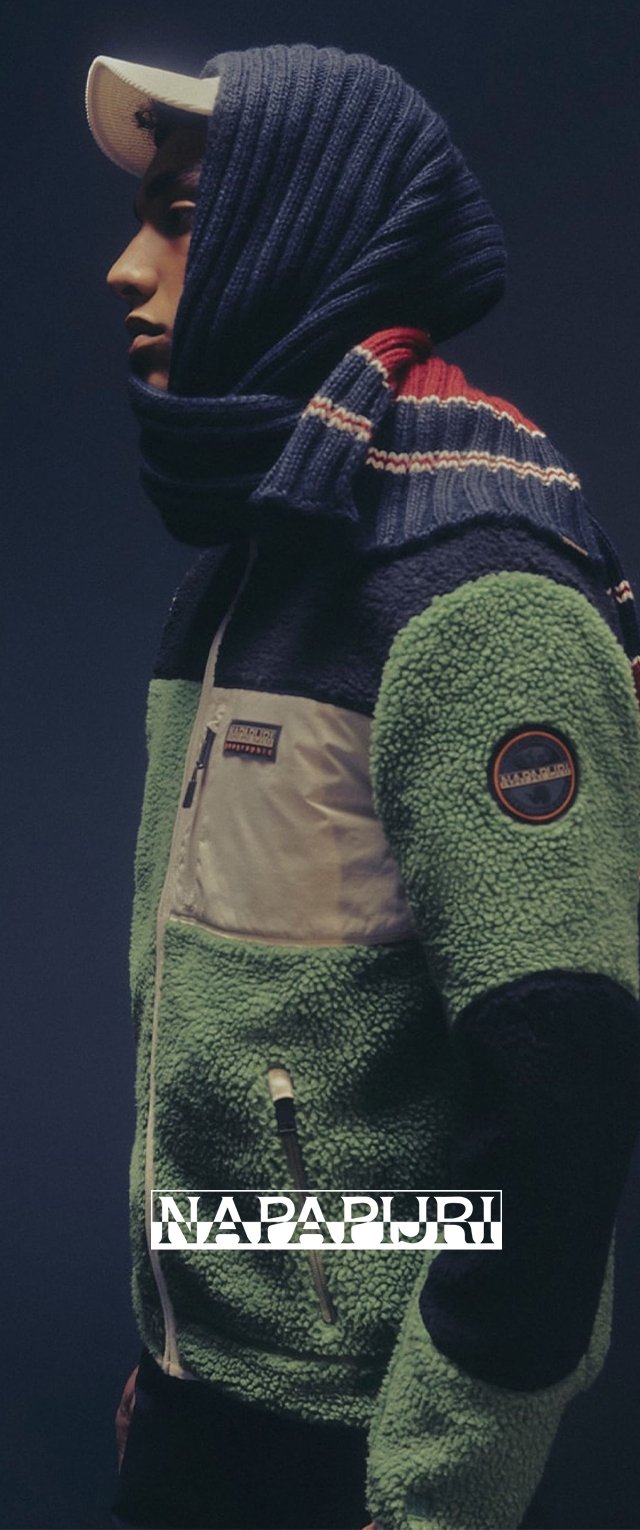 Joven vestido con una chaqueta de forro polar verde, negra y beige con un parche de logo, una gorra blanca y una bufanda a rayas azul marino envuelta alrededor de su cabeza.