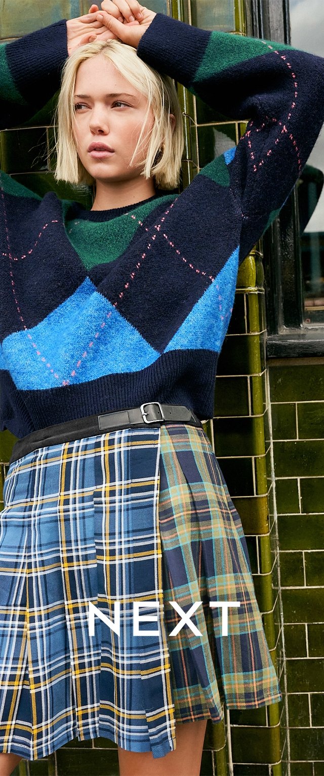 Granatowy sweter w niebiesko-zielony diamentowy wzór, zestawiony z kratkowaną spódnicą w wielu kolorach oraz czarnym paskiem, na tle zielonej, kafelkowej ściany.