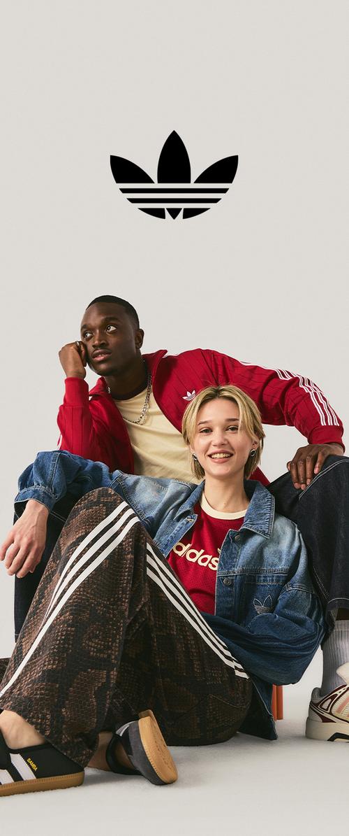 Zwei junge Leute, die Adidas-Kleidung und Schuhe tragen, sitzen lässig unter einem schwarzen Adidas-Trefoil-Logo auf einem schlichten Hintergrund.
