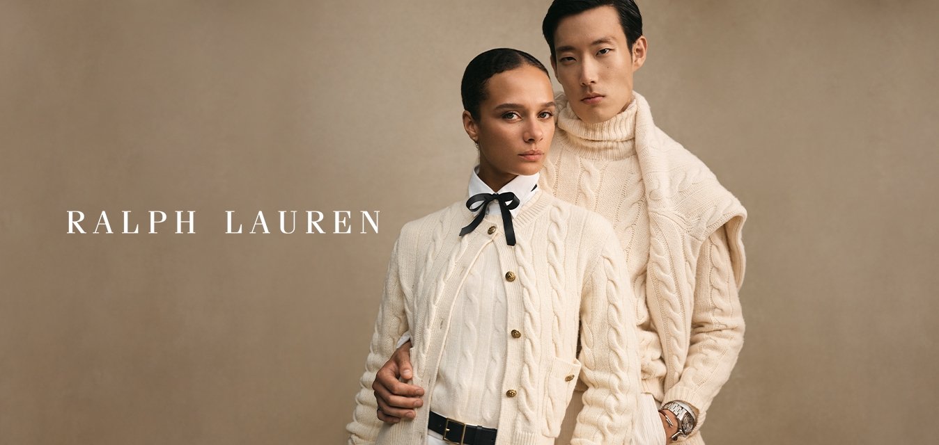 Dva modelky ve smetanových svetroch s pleteným vzorem se úzce postavily proti béžovému pozadí, jedna s motýlkem a druhá s hodinkami.