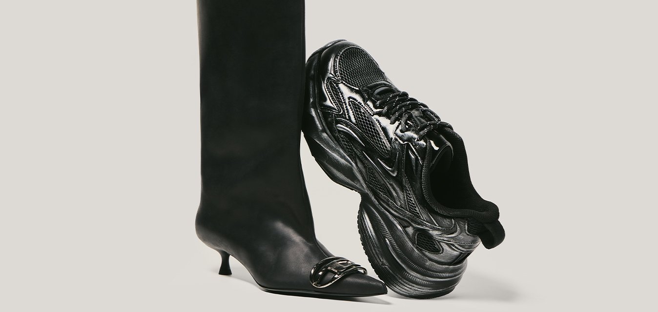 Černá kolenní vysoká kozačka na podpatku s řetízkovým detailem vedle černé masivní atletické tenisky na jednobarevném světlém pozadí.