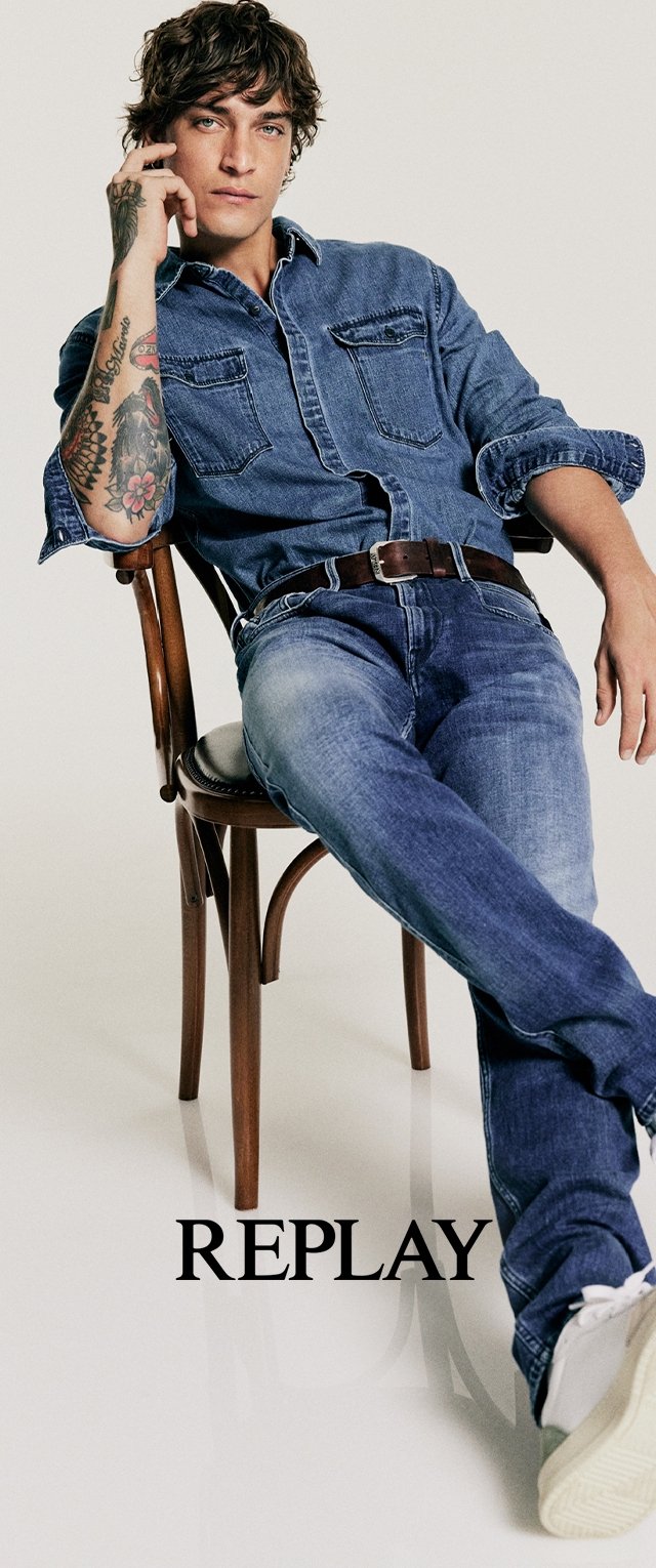 Denimhemd mit Fronttaschen, blaue Jeans, brauer Gürtel, sitzt auf einem Holzstuhl, weiße Sneaker, neutraler Hintergrund, fettgedruckter "REPLAY"-Text darunter.