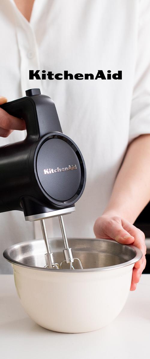 Person hält einen schwarzen KitchenAid Handmixer mit Rührbesen in einer weißen Rührschüssel auf einer weißen Oberfläche.