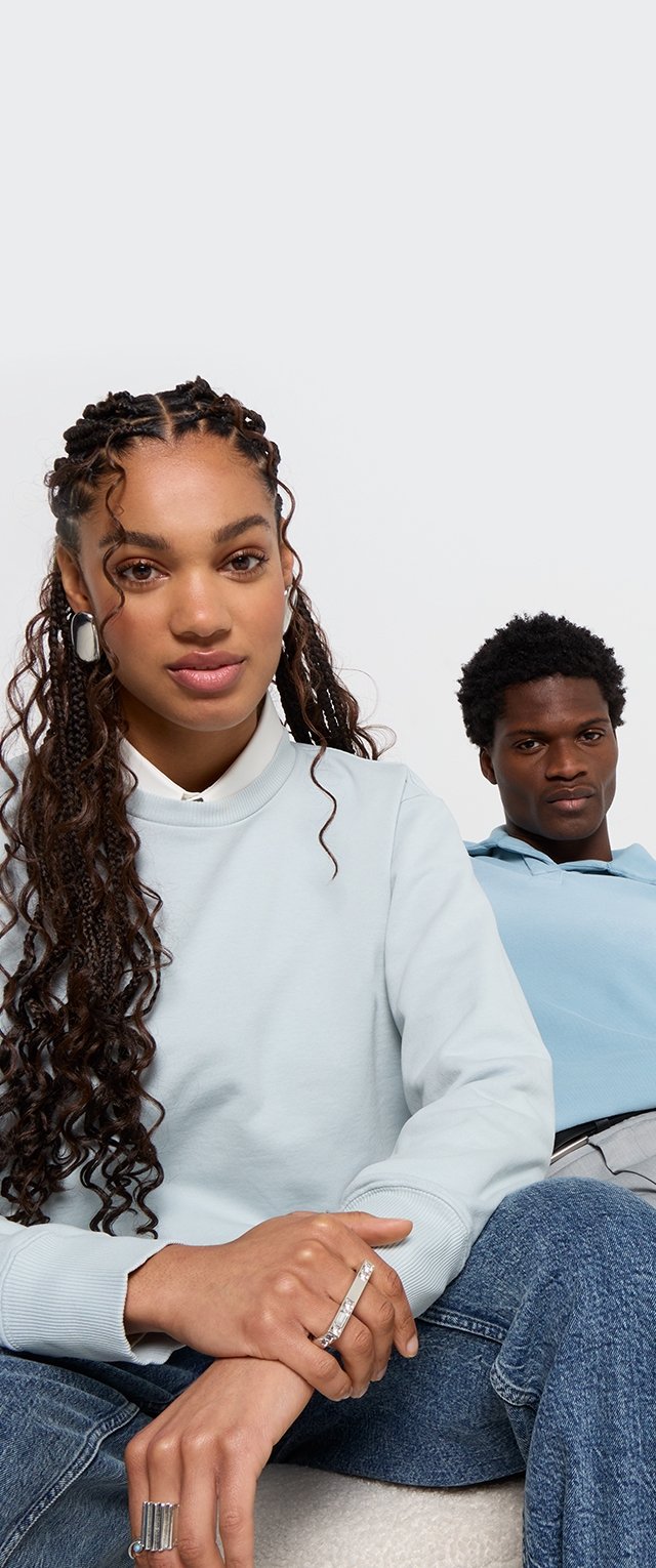 Dos personas están sentadas, vistiendo sudaderas de color azul claro. Una tiene el cabello largo y rizado y lleva joyas; la otra tiene el cabello corto y oscuro y una sudadera con capucha.