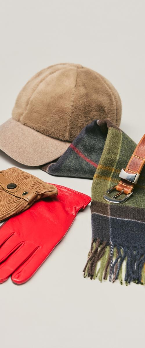 Braune Fleece-Mütze, roter Lederhandschuh und grün kariertes Schal mit Fransen. Die Textur reicht von glattem Leder bis zu weichem Stoff. Schnallendetail am Schal.