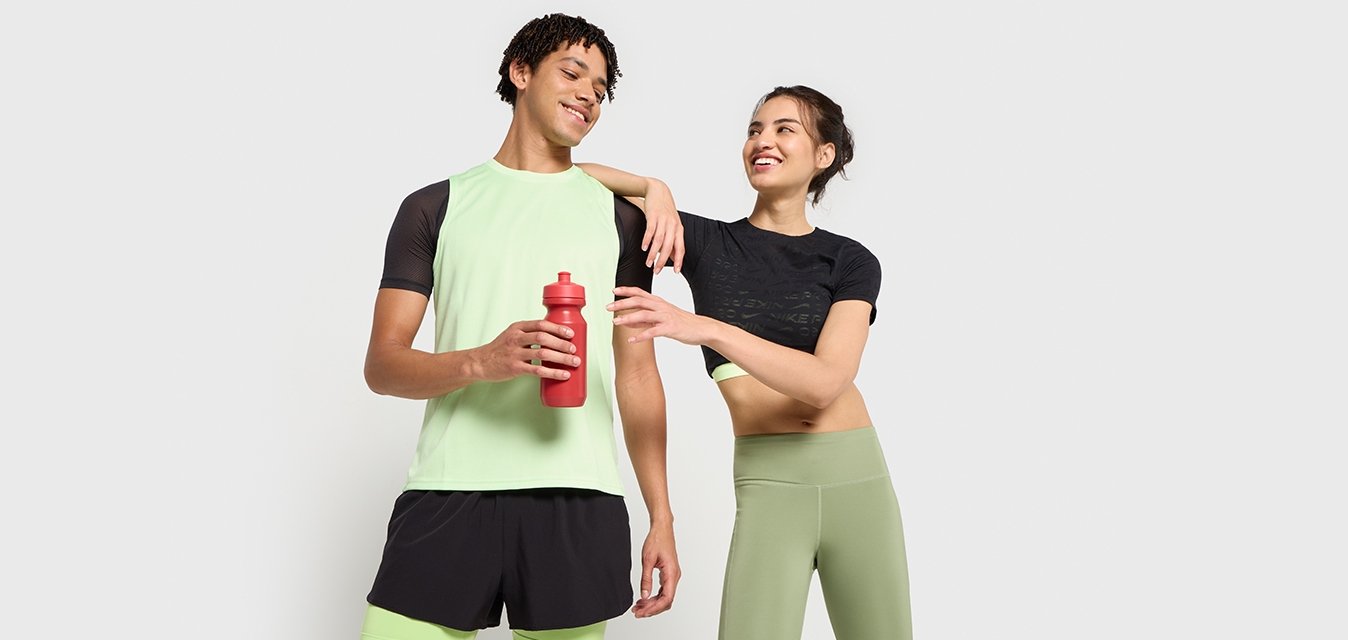 Sportovní oblečení: muž v světle zeleném tričku a černých šortkách, žena v černém crop topu a zelených legínách, držící červenou láhev na vodu.