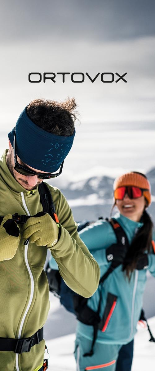 Zwei Personen in Wintersportbekleidung. Der Mann trägt eine enganliegende grüne Jacke mit einem weißen Reißverschluss, ein blaues Stirnband und eine Sonnenbrille. Die Frau hat eine aquafarbene Jacke mit orangefarbenen Akzenten und eine gestrickte orangefarbene Mütze.