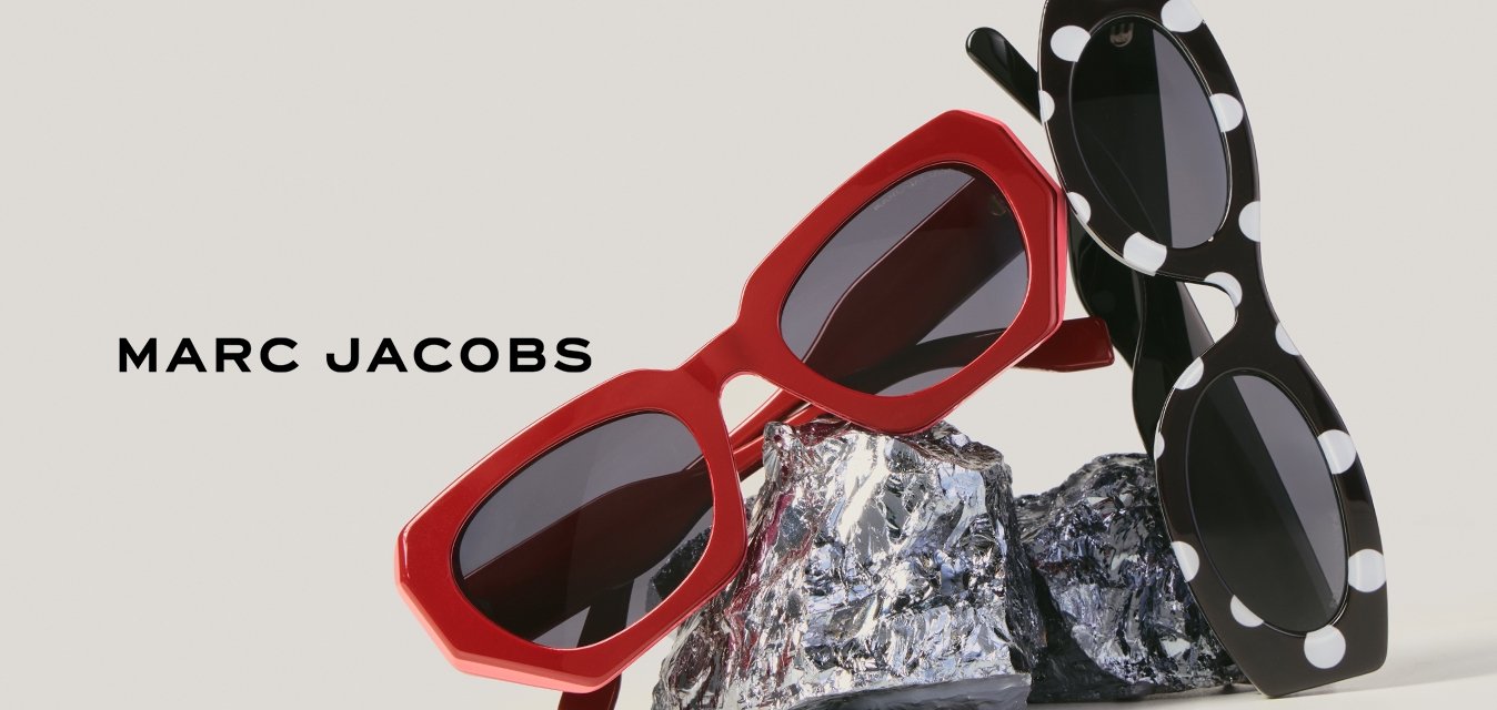 Dva páry slunečních brýlí, jeden červený a druhý černý s bílými puntíky, leží na pomačkané fólii na jednoduchém pozadí.